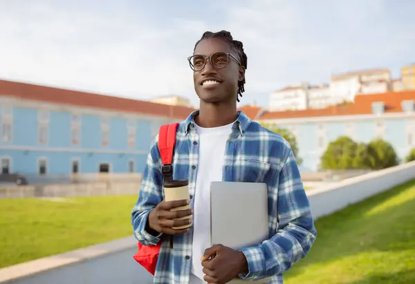 Happy African American college student guy stands holding laptop and coffee cup, blending leisure with learning outside at university park. E-learning and modern digital approach to education