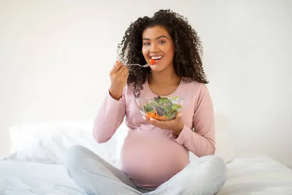 健康な妊娠栄養について 陽気な黒妊娠中の女性は 自宅でベッドに座っている間に新鮮な野菜サラダを食べ カメラで美味しい有機食品と笑顔のボウルを保持し スペースをコピー — ストック写真
