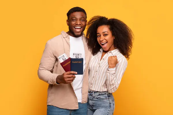 エキサイティングな若い黒人のカップルが搭乗券を持ってパスポートを保持し 勝利のジェスチャーを示し 黄色の背景での彼らの今後の旅行に満足しています — ストック写真