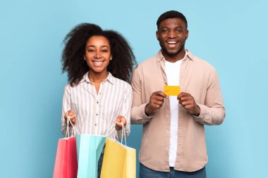 Altın kredi kartı ve renkli alışveriş torbaları sergileyen neşeli genç çift turkuaz arka plana karşı memnuniyet içinde gülümsüyor başarılı alışverişleri ve finansal özgürlüğü sembolize ediyor.