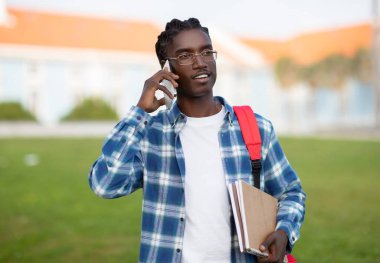 Sırt çantası ve çalışma kitaplarıyla yürürken gülümseyen Afrikalı Amerikalı öğrenci cep telefonuyla konuşuyor neşeli genç adam keyifli telefon konuşmaları ve iletişimi seviyor.