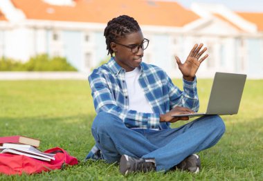 Bilgisayarda el sallayarak ders çalışan mutlu zenci öğrenci. Üniversite kampüsündeki çimlerin üzerinde oturuyor. Video konuşması sırasında ya da uzaktan sanal konferans sırasında e-öğrenmeye dalmış.