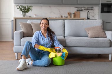 Rahatlamış, gülümseyen bir kadın bir kova temizlik malzemesiyle yerde rahatça oturur, modern oturma odasını toplamaya ara verir,