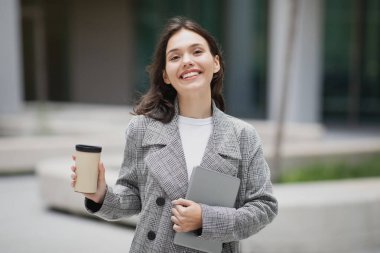 Mutlu genç iş kadını tabletini ve kağıt kahve fincanını elinde tutuyor, şehir ortamında neşeli bir gülümsemeyle kameraya mutlu bir şekilde bakıyor. Çevrimiçi iş ve öğrenim