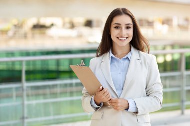 Beyaz tenli, genç, profesyonel bir kadın yönetici elinde bir dosya, temiz bir bej takım elbise ve mavi gömlek, kurumsal bir ortamda güven ve verimliliği temsil ediyor.