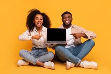 Yerde bağdaş kurmuş oturan mutlu siyah erkek ve kadın, sarı arka planda boş bir ekranla mutlu bir şekilde dizüstü bilgisayarı işaret ediyorlar.