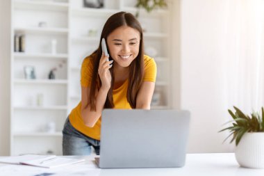 Cep telefonu ve dizüstü bilgisayarlı genç bir Asyalı kadın İçişleri Bakanlığı 'ndan uzaktan çalışıyor, sırıtan Koreli serbest çalışan bir kadın odada duruyor, bilgisayar ekranına bakıyor ve cep telefonuyla konuşuyor.