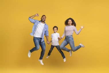 Stüdyoda sarı arka planda zıplayan üç kişilik Afro-Amerikan ailesinin neşeli portresi mutlu siyahi ebeveynler ve küçük oğlu birlikte eğleniyor.