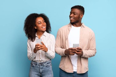 Her biri kendi akıllı telefonlarını kullanan mutlu genç siyah çift, bakışlarını ve gülümsemelerini, yumuşak mavi arka plana karşı