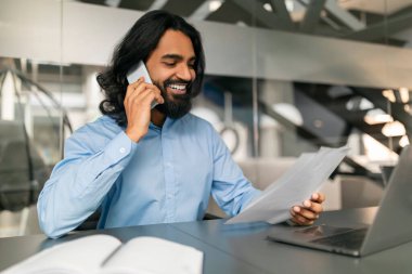 Ofiste çalışan neşeli Hintli genç Hintli CEO yöneticisi, masada oturuyor, telefon görüşmeleri yapıyor, dizüstü bilgisayar kullanıyor, gazete pazarlama raporlarını kontrol ediyor, fotokopi çekiyor. Başarılı kariyer konsepti