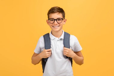 Gözlüklü ve sırt çantalı neşeli bir öğrenci sarı stüdyo geçmişine güvenle bakıyor. Okula hazır, öğrenmenin ve eğitimin tadını çıkaran mutlu bir genç çocuk.