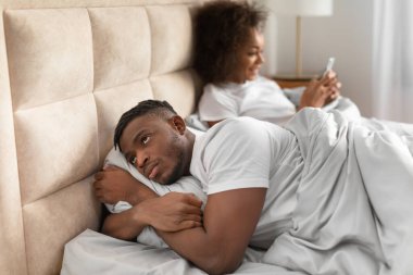 Infidelity, jealousy issue. Jealous black husband discontented about wifes online chats while she texting on cellphone, man suspecting cheating lying in bed in domestic bedroom. Selective focus clipart