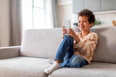 Zeki telefon ekranına gülen neşeli siyah çocuk, mutlu Afro-Amerikan ergen erkek çocuk oturma odasında oturmuş video konuşmasından zevk alıyor ya da cep telefonuyla oyun oynuyor.