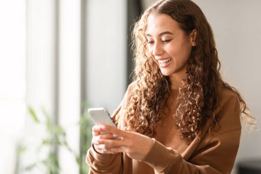 Mutlu genç kız öğrenci telefonuyla interneti tarıyor, evde oturuyor, rahat modern yaşam tarzını ve dijital teknoloji kullanımını somutlaştırıyor. Hafta sonları arkadaşlarıyla mesajlaşan genç bayan.