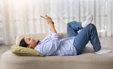 Telekomünikasyon, mobil uygulama konsepti. Milenyum Asyalı neşeli kadın rahat kıyafetler giyip evdeki rahat oturma odasında kanepede uzanıyor, akıllı telefon kullanıyor, video çekiyor, yan görüş alıyor.