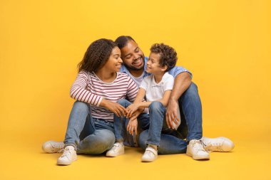 Mutlu Afrikalı genç Amerikalı ebeveynler, küçük oğullarıyla yerde oturuyor, erkek çocuğa sarılıyorlar ve birbirlerine gülümsüyorlar, üç çocuklu neşeli zenci aile sarı stüdyo arka planında poz veriyorlar.