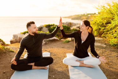 Erkek ve kadın yoga minderlerine oturmuş, sabahın ilk ışıklarıyla deniz kenarında huzurlu yoga antrenmanının ortasında beşlik çakıyor ve gülümsüyor.