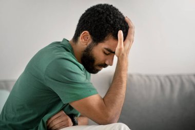 Üzüntülü, sakallı, yeşil tişörtlü, gri bir kanepede oturan, kafasını elinde tutarken endişeli yüzünü ifade eden orta doğulu genç bir adam.