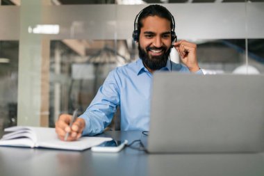 Uzaktan iletişim konsepti. Kulaklıklı neşeli Hintli adam bilgisayarda çalışıyor, ofiste video görüşmesi yapıyor. Milenyum Doğulu adam web kamerasından konuşuyor, iş görüşmesi yapıyor.