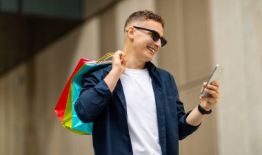 Şık, mutlu, güneş gözlüklü bir beyaz adam, akıllı telefonuna bakıyor, omuzunda renkli alışveriş torbaları taşıyor, koyu renk bir ceket ve şık bir ayakkabı giyiyor.