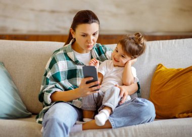 İnternetten alışveriş. Sevimli küçük kız ve annesi evde akıllı telefon kullanıyor, mutlu genç anne ve okul öncesi kızı cep telefonuyla koltukta dinleniyor, internetten satın almanın tadını çıkarıyor.