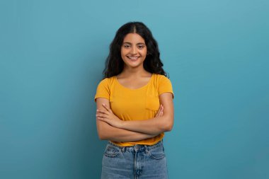 Sarı tişörtlü, mavi arka planda poz veren ve kameraya gülümseyen kıvırcık saçlı, neşeli, genç Hintli kadının stüdyo çekimi. Milenyum yaşam tarzı