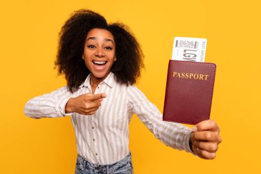 Neşeli genç siyahi bayan gezgin elinde bordo pasaportuyla geziyor ve neşeli sarı arka planda tatil için hazır olduğunu ifade ediyor.
