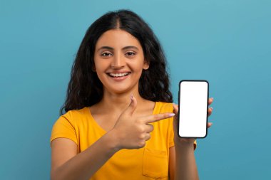 Heyecanlı, neşeli, sarı tişörtlü, beyaz ekranlı büyük bir telefonu işaret eden, mobil uygulama, online teklif, mavi arka plan, maket, kopyalama alanı gösteren genç doğu kadın öğrenci.