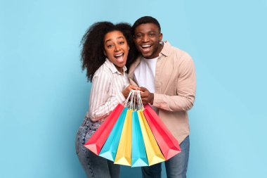 Afro-Amerikalı parlak genç çift, neşe dolu alışveriş torbalarıyla gülüyor, ferahlatıcı mavi arka plana karşı.