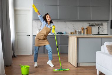 Sarı eldivenli, neşeli genç bir kadın yeşil paspası geçici mikrofon olarak kullanıyor, ahşap zeminli, neşeli bir şekilde mutfakta şarkı söylüyor.