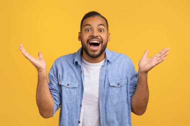 Şaşırmış genç Afrikalı Amerikalı adam kameraya ağzı açık bakıyor ve ellerini kaldırıyor, duygusal siyah erkek sarı stüdyo arka planında el kol hareketi yapıyor, hayret verici, yakın çekim