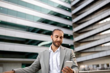 Takım elbiseli, akıllı telefonuyla meşgul olan, arka planda modern ofis binasına karşı duran, yakışıklı erkek girişimci mesajlaşması, bağlantıyı sembolize ediyor.
