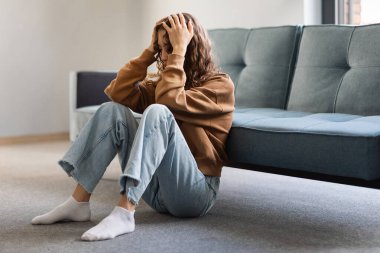 Genç bayan üzgün ve depresif bir şekilde evde oturuyor, umutsuzluk içinde kafaya dokunuyor, depresyonla boğuşuyor ve zihinsel problemlerle boğuşuyor, oturma odasında yerde oturuyor. Boş boşluk