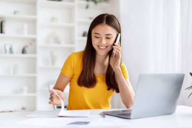 Planlama Konsepti. Ev ofisindeki masada çalışan mutlu genç Asyalı kadın, gülümseyen Koreli kadın cep telefonuyla konuşuyor ve not defterine bakıyor, notları ve çalışma saatlerini kontrol ediyor, boşluğu kopyalıyor.