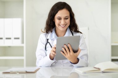 Modern sağlık hizmetleri, tıp teknisyenleri. Gülümseyen Avrupalı doktor masasında oturuyor, klinik hastanesinde dijital tablet kullanıyor, hastaya internetten mesaj atıyor, reçete yazıyor.