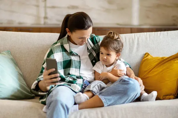 自宅でスマートフォンで小さな娘にオンラインコンテンツを表示する母親は リビングルームのインテリアでソファーに座って ママと一緒に漫画を見ながら飛んでいるトッドラーの少女を怒らせる — ストック写真