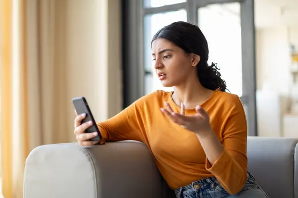 沮丧的印度年轻女人带着怀疑的表情看着她的智能手机 手拿着手机坐在客厅的沙发上 阅读着令人不快的信息 — 图库照片