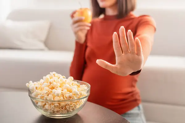 No Junk Food During Pregnancy. Unrecognizable Pregnant Woman Refusing To Take Unhealthy Popcorn Snack Choosing Healthy Apple Fruit, Sitting On Sofa In Living Room Indoor. Cropped, Selective Focus