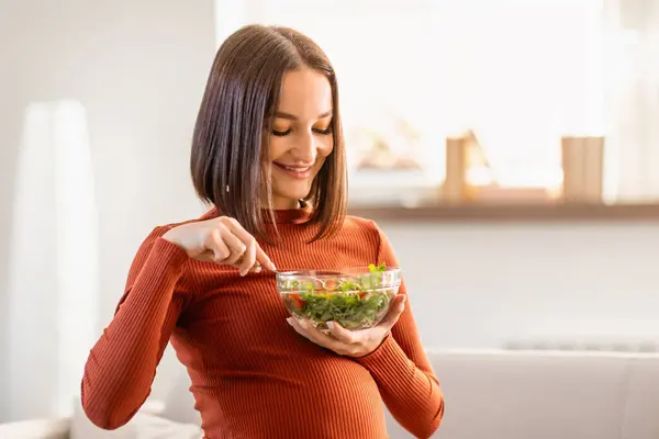 Healthy diet in pregnancy. Happy pregnant young woman eating healthy fresh salad at home. Cheerful expecting European lady enjoying vegetable meal while sitting on sofa indoors