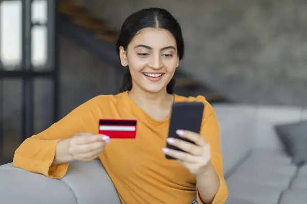 スマートフォンとクレジットカードを使用して笑顔のインドの女性 オンラインショッピング 快適にスタイリッシュなインテリアでソファに座って インターネットで購入し 支払いを行う — ストック写真