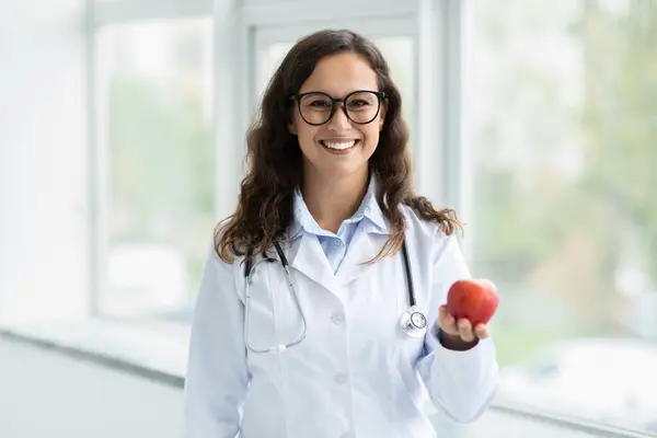年轻美丽的欧洲裔女性营养学家戴着眼镜 手里拿着苹果 推荐新鲜水果 以促进健康饮食和营养 站在诊所窗边 复制空间 — 图库照片