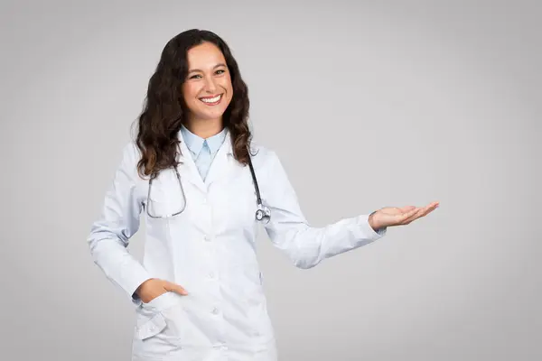 白いコートとステスコープの自信を持った若い女性医師は笑顔で 灰色の背景 自由なスペースに対して片手でプレゼンテーションジェスチャーを作ります — ストック写真