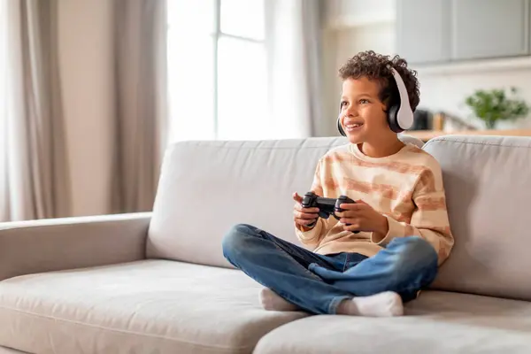 自宅でゲームコントローラーを使用してヘッドフォンに巻き込まれた黒人の少年 リビングルームのインテリアで快適なソファーでリラックスしながらビデオゲームを楽しんでいる幸せな19歳の男性の子供 コピースペース — ストック写真