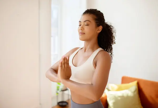 Sporty lady meditating with hands clasped standing in the calm of her living room, embodying mindfulness and balance in her morning yoga practice, posing with eyes closed