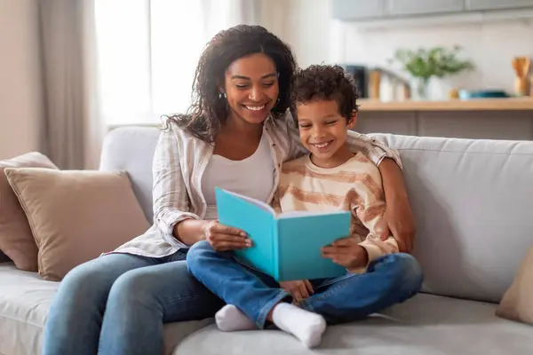Cheerful black mother and her preteen son reading book together at home, happy african american mom and male kid engrossed in storybook while relaxing on comfortable couch in living room