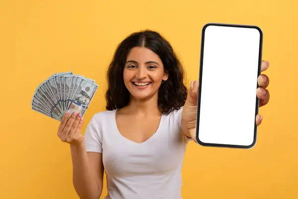 幸せな美しい若いインドの女性ギャンブルインターネット上で 白い空の画面を持つスマートフォンを使用して ドル現金 黄色のスタジオの背景 モックアップの束を示しています オンラインベットコンセプト — ストック写真