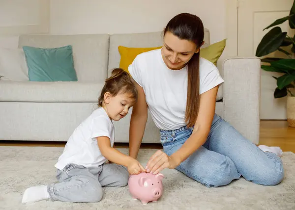 Happy Family Mother And Little Daughter Putting Coins In Piggy Bank At Home, Mom And Cute Preschool Female Child Saving Money For Future, Enjoying Economy While Relaxing In Living Room Together