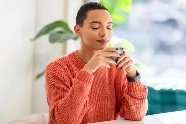 Relaxed latin young woman enjoys the aroma of her drink, eyes closed, wearing a cozy orange sweater and a smartwatch in a bright, plant-filled room. Lifestyle alone, free time