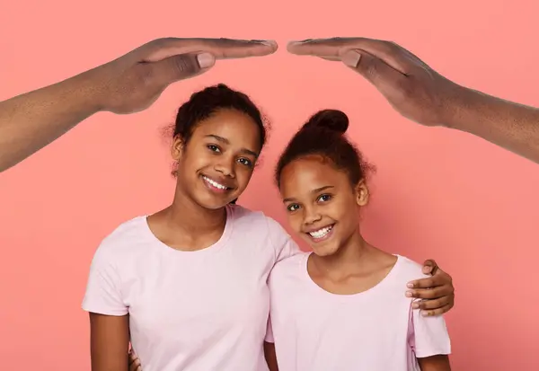 Cute smiling african american girls sisters teenagers wearing pink t-shirts hugging under hands roof, colorful studio background, collage. Child safety concept, insurance for kids