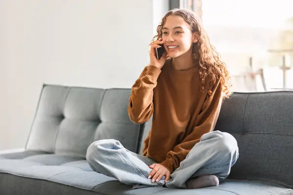 Young teen lady speaking on mobile phone, having cheerful communication with friend, sitting on sofa at home interior. European teenager girl smiling as she chats on smartphone gadget